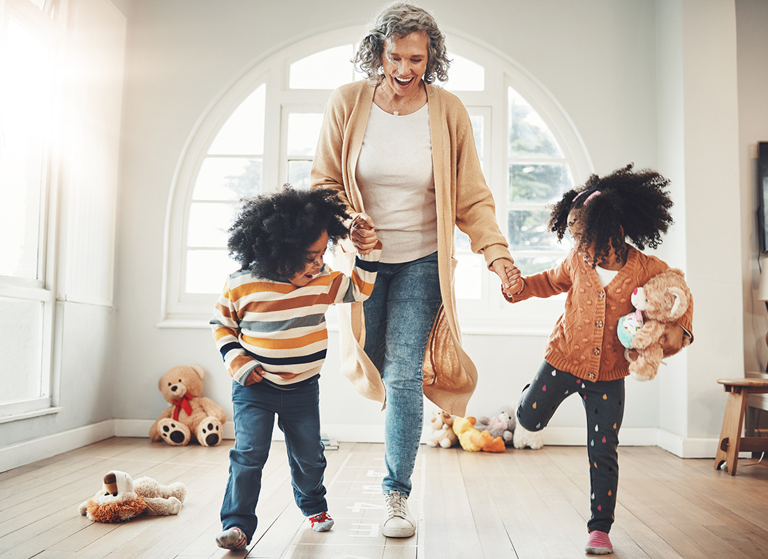 About Our Agency - View of a Cheerful Grandmother Having Fun Playing Hopscotch While Holding Hands with her Two Granddaughters at Home