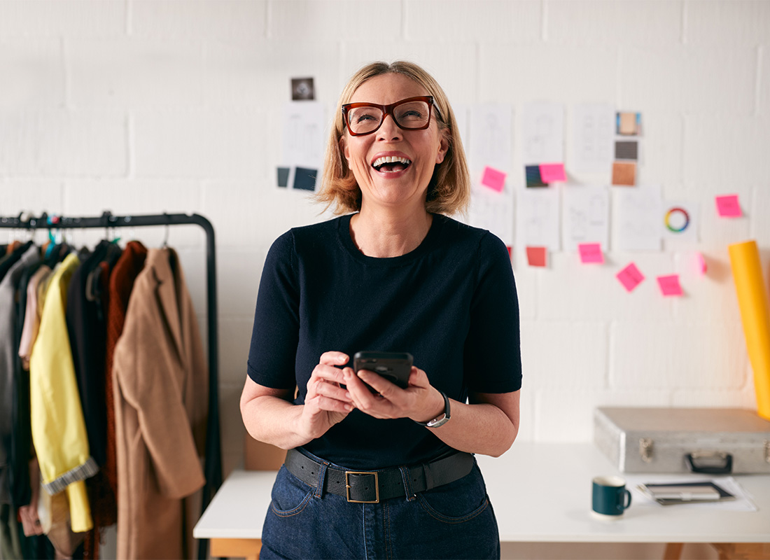 Service Center - Closeup Portrait of a Cheerful Young Small Business Owner Standing in her Clothing Studio While Holding a Phone in her Hands