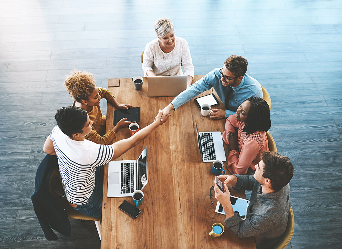 We Are Independent - Small Group of Employees Sitting Around a Wooden Table in a Conference Room While Two People Shake Hands Across the Table During a Meeting