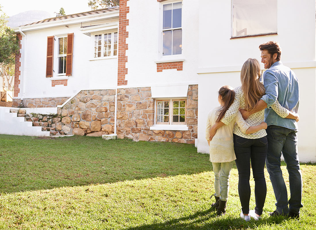 Personal Insurance - Rear View of a Family of Three with a Young Daughter Standing Outside Their New Home on the Green Grass on a Sunny Day